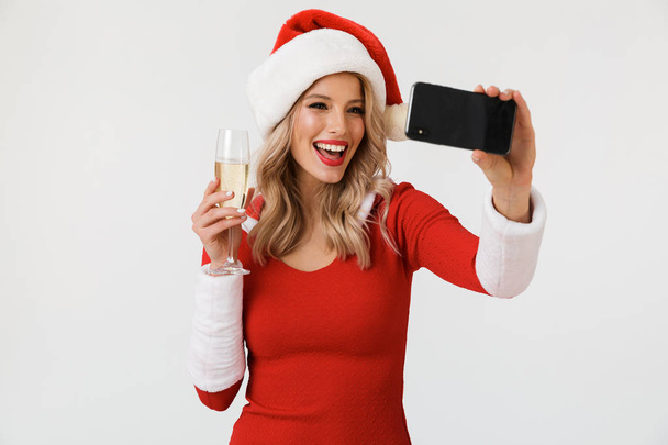 Portrait of a smiling blonde woman dressed in red New Year costume standing isolated over white background, holding glass of champagne, celebrating, taking a selfie - Zdjęcie, obraz
