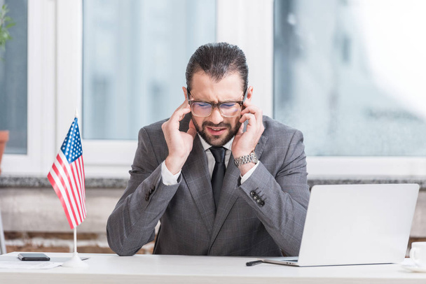 разочарованный бизнесмен сидит за столом офиса с ноутбуком и американским флагом
 - Фото, изображение