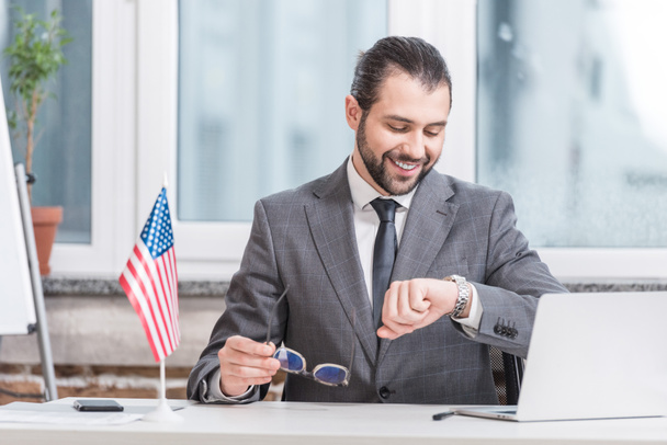smiling businessman sitting at table with laptop and little american flag and looking at watch - Foto, afbeelding
