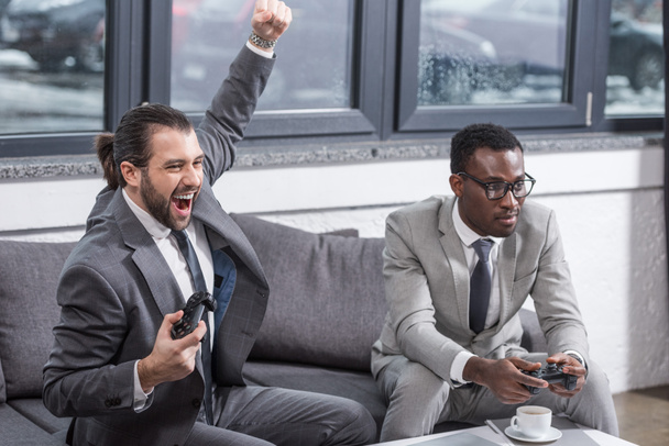 handsome businessman holding gamepad, raising hand and screaming while african american partner looks like loser - Foto, Imagem