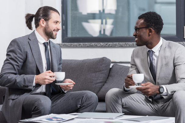 erfolgreiche multiethnische Geschäftsleute, die im Büro auf der Couch sitzen und Kaffee trinken - Foto, Bild