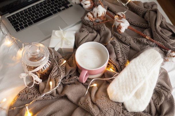 Mug of hot cappuccino on the bed. Cozy decor. Breakfast. Mug, plaid, cotton, laptop, candle. Gift box and knitted mittens. Christmas lights. Holidays. - Photo, Image