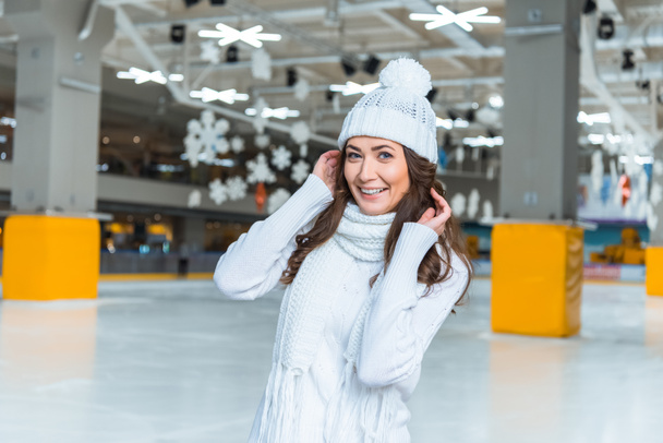portrait of smiling beautiful woman in hat and sweater looking at camera on skating rink - Foto, Imagen