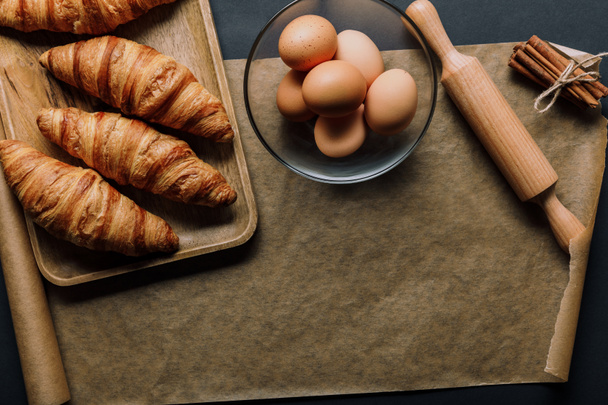 elevated view of  cinnamon, eggs in bowl, tray with croissants, rolling pin and baking paper on black table - Fotó, kép