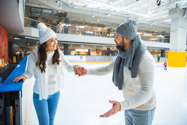 smiling couple in hats and sweaters holding hands while skating on ice rink - Photo, Image