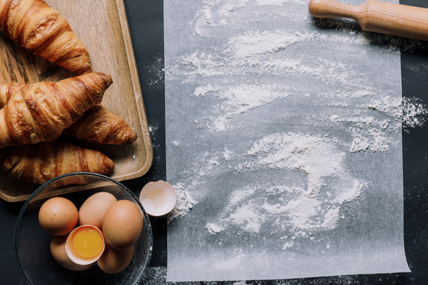pohled shora z mouky na pečící papír a croissanty s vejci v misce na černý stůl  - Fotografie, Obrázek