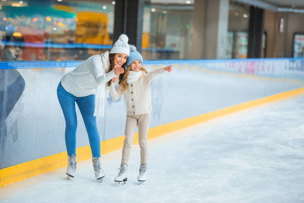 smiling mother and daughter pointing away on skating rink - Photo, Image