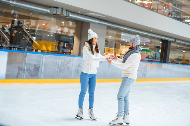 smiling couple in hats and sweaters holding hands while skating on ice rink - Photo, Image