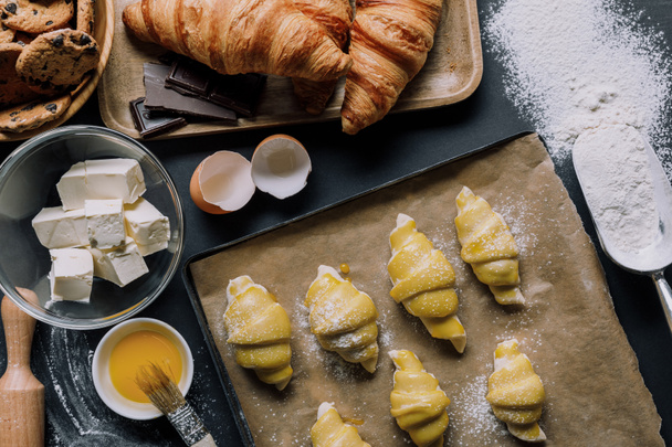 flat lay with dough for croissants on tray, yolk with brush and ingredients on table covered by flour - Fotó, kép