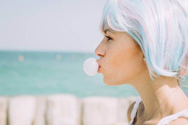 joven mujer en rosa y azul peluca soplando un chicle en th playa
 - Foto, Imagen
