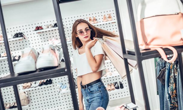 Portrait of a smiling woman in sunglasses holding shopping bags on storefront with shoes background. - 写真・画像