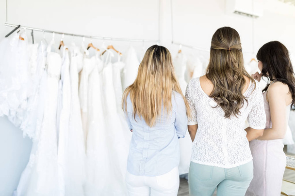 Вид сзади запутавшихся друзей, смотрящих на свадебные платья, висящие в бутике
 - Фото, изображение