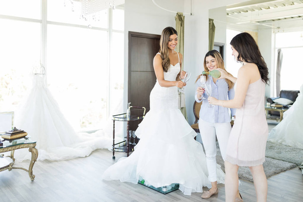 Vrouwelijke koper in trouwjurk met champagne met beste maatjes in bruids boutique - Foto, afbeelding