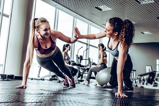 Two attractive fitness girls doing push ups. Girls give five each other. Gym background. - Valokuva, kuva