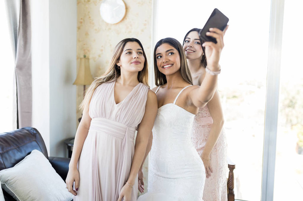 Smiling young bride and bridesmaids taking selfportrait on wedding day in living room - Photo, Image
