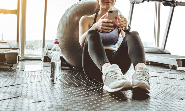 Young woman athlete using cell phone at gym. Young woman in sportswear checking phone while resting after workout on floor. Beautiful fit girl messaging with smartphone at fitness centre. - Foto, imagen