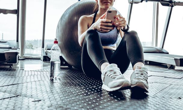 Young woman is sitting on mat near bottle of water and looking at phone while having rest after training in gym. - Fotó, kép