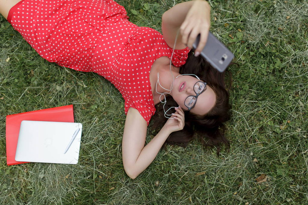 Beautiful woman takes selfie lying on the grass surrounded her papers. Top view - Foto, Imagen