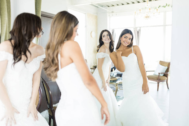 Cute young brides in white dresses looking into mirror at wedding boutique - Фото, изображение