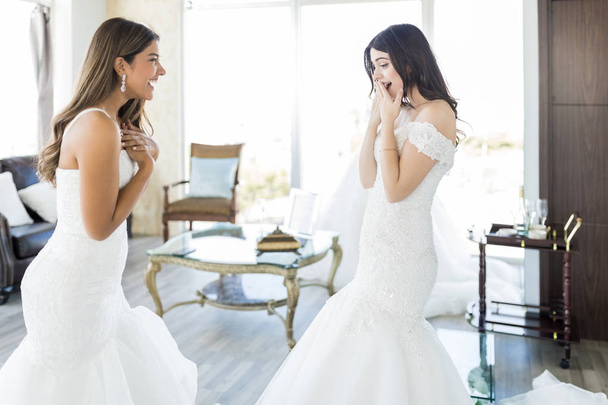 Cheerful and astonished young women in elegant wedding gowns at bridal shop - Foto, imagen