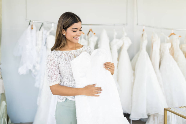 Cute Hispanic woman choosing white wedding outfit in bridal shop - Фото, изображение