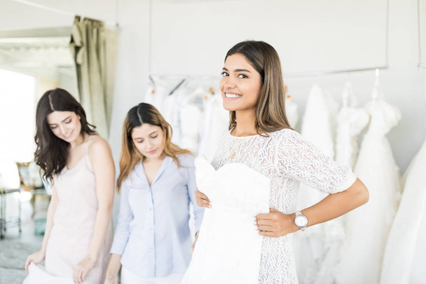 Smiling Latin woman holding her perfect wedding dress with friends in clothing store - Photo, Image