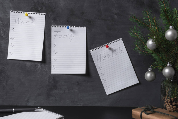 Офисный стол с рождественскими украшениями и страницы блокнота прикреплены к стене, с контрольными листами, касающимися работы, семьи и здоровья. Концепция планирования
. - Фото, изображение