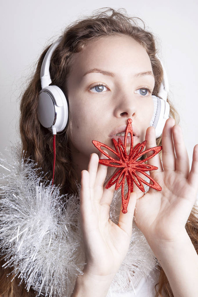 smiling redhead woman in white scarf listening music with headphones in front of white wall and holding red decorative snowflake, christmas concept - Photo, Image