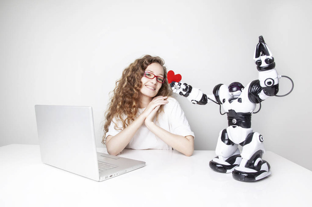 smiling redhead woman working with laptop in front of white wall while robot giving red heart to her - Photo, Image