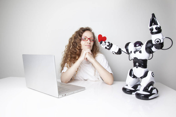 attractive redhead woman working with laptop in front of white wall while robot giving red heart to her - Photo, Image