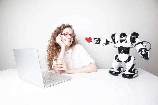 счастливая рыжая женщина работает с ноутбуком перед белой стеной, в то время как робот дает ей красное сердце
 - Фото, изображение