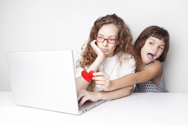 overwerkt roodharige vrouw die werkt met laptop voor witte muur terwijl haar dochter om haar rood hart van achteren - Foto, afbeelding