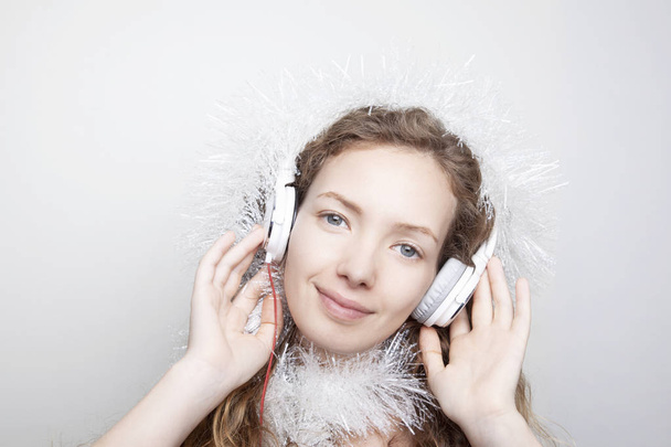 mujer pelirroja sonriente en bufanda blanca escuchando música con auriculares frente a la pared blanca y mirando a la cámara
 - Foto, Imagen