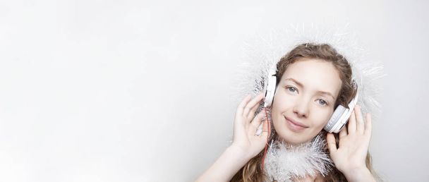 χαμογελώντας Κοκκινομάλλης γυναίκα στο λευκό μαντήλι ακούγοντας μουσική με ακουστικά μπροστά από το λευκό τοίχο, και βλέπουν φωτογραφική μηχανή - Φωτογραφία, εικόνα