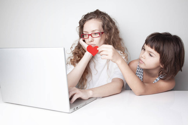 Müde rothaarige Frau arbeitet mit Laptop vor weißer Wand, während ihre Tochter ihr rotes Herz von hinten schenkt - Foto, Bild