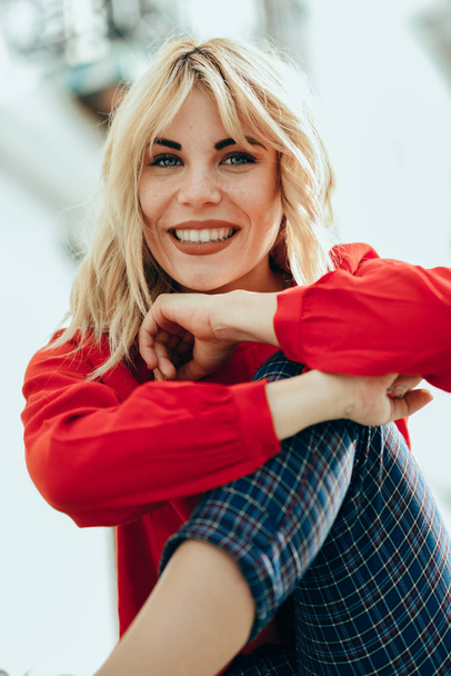 Happy young blond woman sitting on urban background. Smiling blonde girl with red shirt enjoying life outdoors. - Fotó, kép