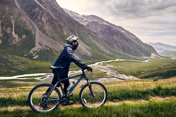 Man on mountain bike in helmet in the mountain valley with river at cloudy sky background. - Photo, Image