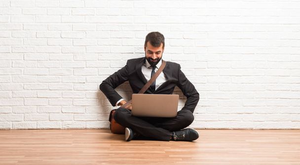 Uomo d'affari con il suo computer portatile seduto sul pavimento in posa con le braccia all'anca e ridendo guardando davanti
 - Foto, immagini