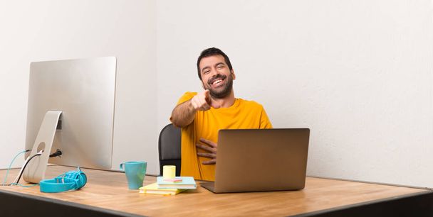 Чоловік працює з ноутбуком в офісі, вказуючи пальцем на когось і багато сміється
 - Фото, зображення