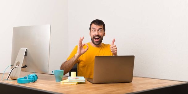 Homem trabalhando com laptot em um escritório mostrando sinal ok com e dando um gesto polegar para cima
 - Foto, Imagem