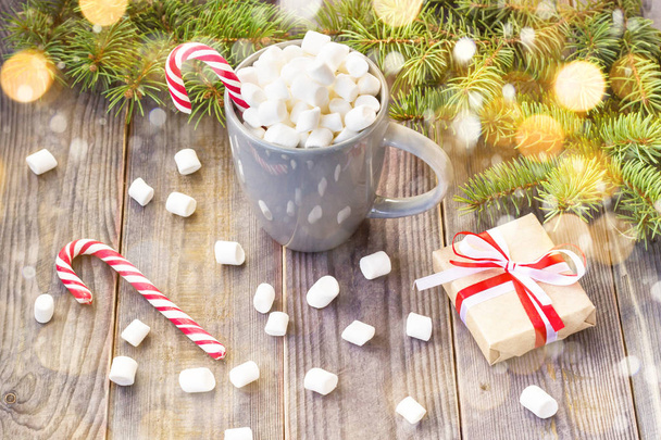 Warme koffie chocolade met marshmallow in kerstsfeer, feestelijke decoratie op rustieke houten tafel achtergrond, candy canes geschenk dozen rood lint fir boom versierd met sneeuw - Foto, afbeelding