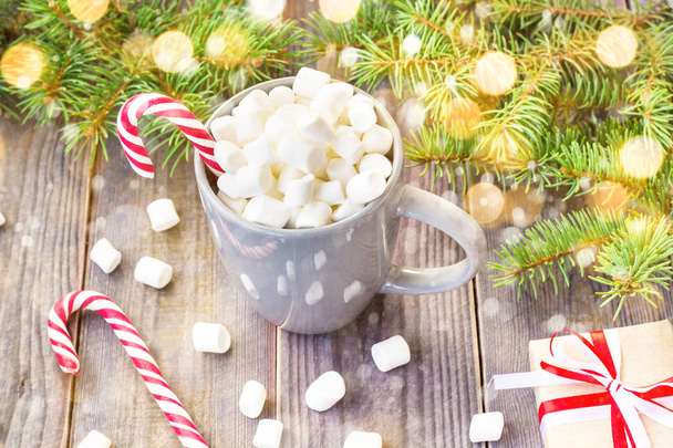 Warme koffie chocolade met marshmallow in kerstsfeer, feestelijke decoratie op rustieke houten tafel achtergrond, candy canes geschenk dozen rood lint fir boom versierd met sneeuw - Foto, afbeelding