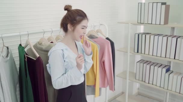 Asian fashion female blogger online influencer holding shopping bags and lots of clothes on clothes rack for recording new fashion video broadcast live video to social network by internet at home. - Footage, Video