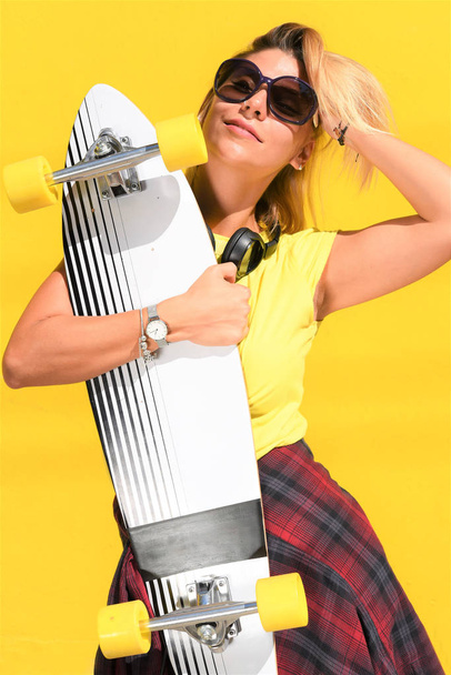 красивая молодая женщина со скейтбордом и солнечными очками на желтом фоне - Фото, изображение