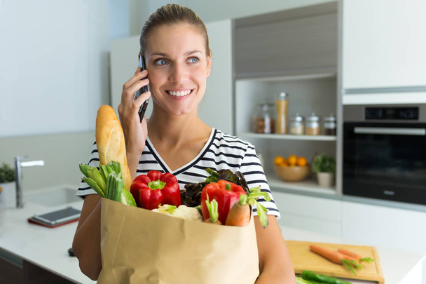 自宅の台所で食料品の袋で野菜を保持しながら彼女の電話で話している健康な若い女性のショット. - 写真・画像