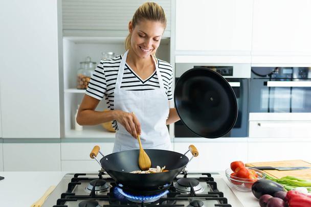Laukaus terve nuori nainen ruoanlaitto ja sekoittamalla ruokaa paistinpannu keittiössä kotona
. - Valokuva, kuva
