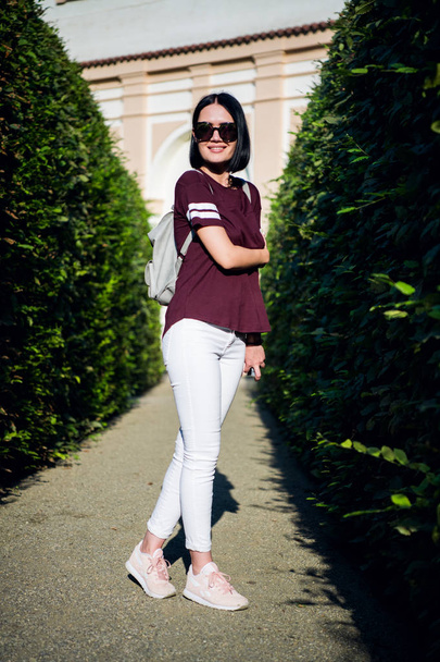 Close-up lifestyle fashion portrait of young hipster woman walking at park , travel with backpack, stylish casual outfit, evening sunset, reflecting sunglasses student - Photo, Image