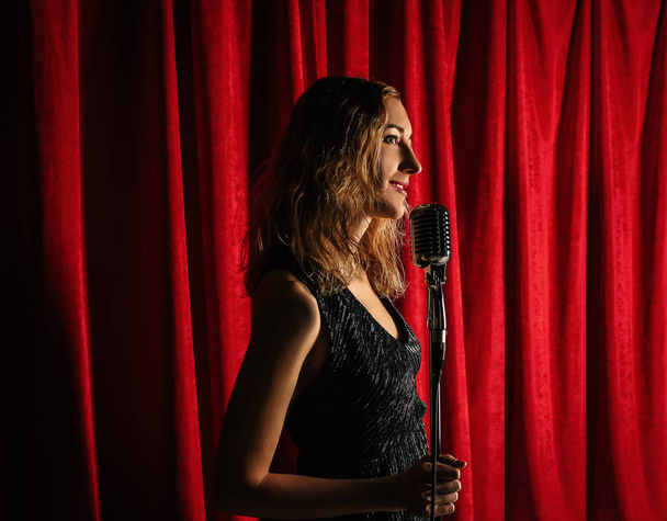 Young attractive girl singing on stage with microphone against the background of red curtains - Foto, Imagem