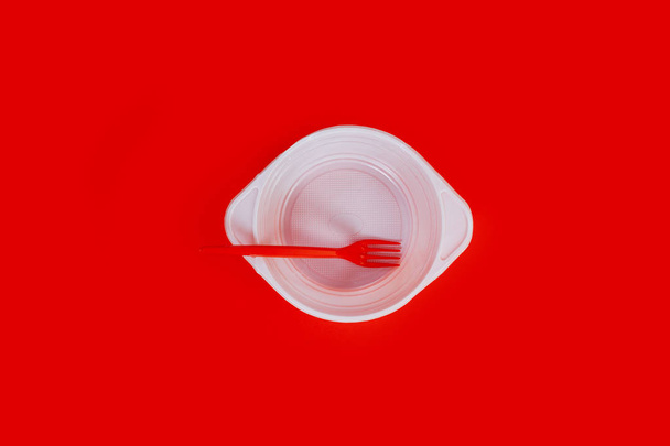 Λευκό πλαστικό πιάτο και κόκκινο πιρούνι σε κόκκινο φόντο. - Φωτογραφία, εικόνα