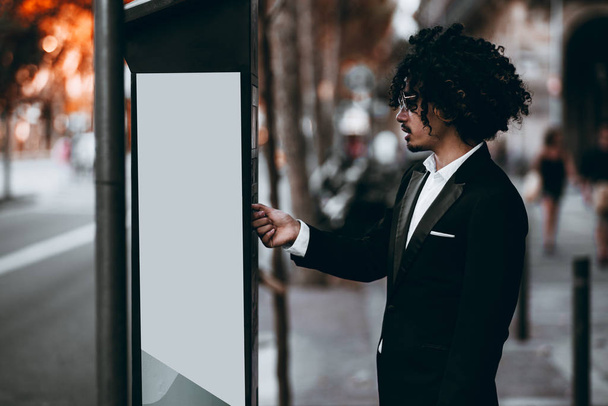 An Asian businessman with curly hair and in sunglasses is paying his parking time via an outdoor terminal; a young man entrepreneur in a formal suit is making payment using parking meter on the street - Fotoğraf, Görsel
